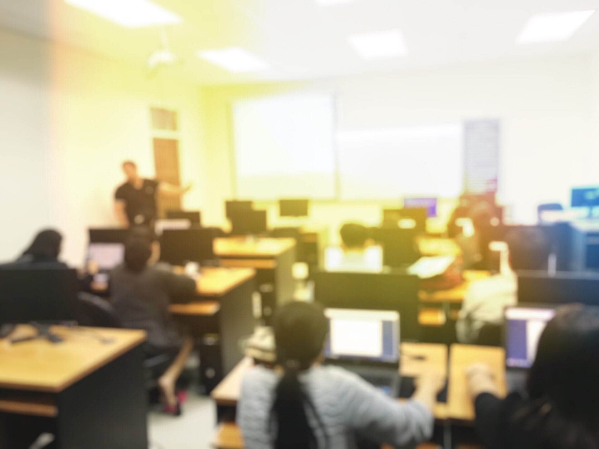 blurred image of student with teacher learning business technology desktop computer in a classroom and screen projector for present. education or training concept